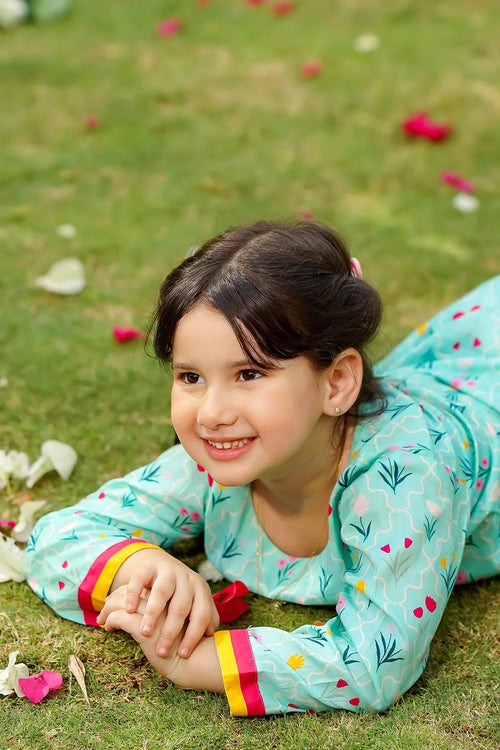Smiling girl in floral mint green dress lying on grass with flower petals, kids fashion outfit