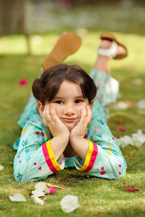 Little girl in floral print dress lying on grass, smiling with hands on cheeks, wearing sandals