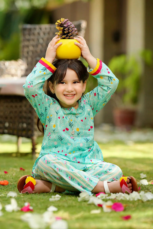 Smiling girl in floral mint green kids outfit, sitting on grass with flower petals, holding yellow bowl with pinecone