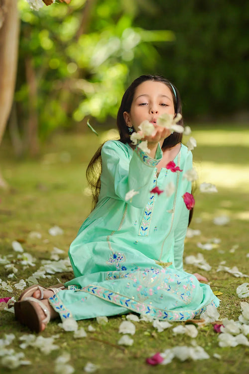 Girl in mint green embroidered dress blowing flower petals, sitting on grass outdoors