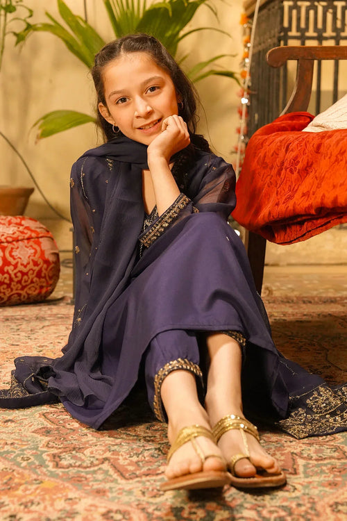 Girl in navy blue embroidered kids outfit with gold sandals, sitting on patterned rug